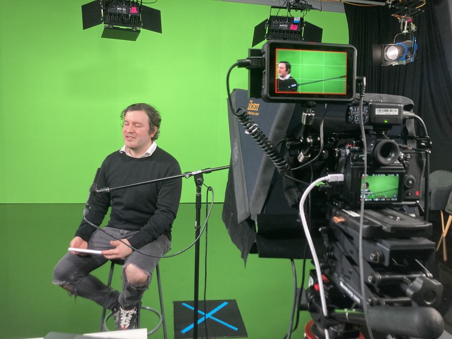 Speaker on a stool in front of a green screen, cinema camera in the foreground