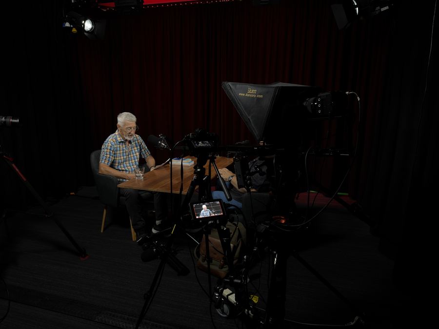 Interviewee at a table lit against a black backdrop, teleprompter camera